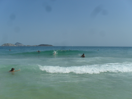 Mar de Ipanema, em uma cor semelhante a turquesa, com uma onda e três banhistas. Céu azul limpo e uma ilha no horizone.