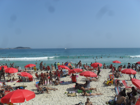 Uma praia com areia branca e mar azul ao fundo. Vários guarda-sóis vermelhos e pessoas na areia.